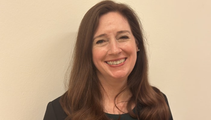 Jennifer Baker with long brown hair and a black shirt smiling in front of a plain beige background.