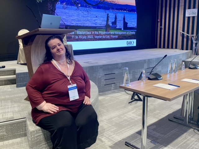 A lady sits infront of a lectern there is a presentation on a large screen in the background