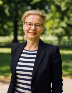 A smiling woman with glasses wearing a striped top and blazer standing outdoors.