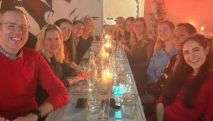 A group of smiling people gathered around a dinner table with a warm, reddish ambiance.