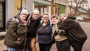 A group of five adults smiling and posing closely together on an urban street.