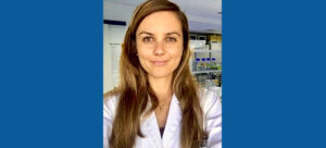 Woman in lab coat smiling at the camera in a laboratory setting with shelves holding lab equipment visible in the background.