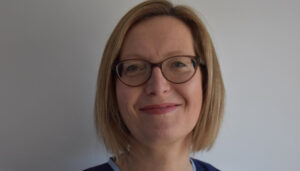 Middle-aged caucasian woman with short blond hair and glasses, smiling at the camera against a plain white background.