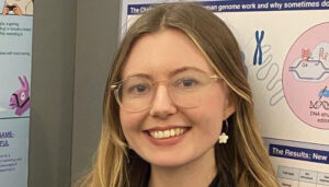 Young woman with glasses smiling at a science exhibition, standing in front of educational posters.