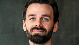 Portrait of a smiling man with a beard and mustache, looking directly at the camera, against a blurred gray background.