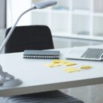 A minimalist office desk features an open laptop, a notebook, sticky notes, a small potted plant, and an adjustable desk lamp.