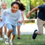 A group of children is running and playing outside on a grassy area. One boy is holding a football.