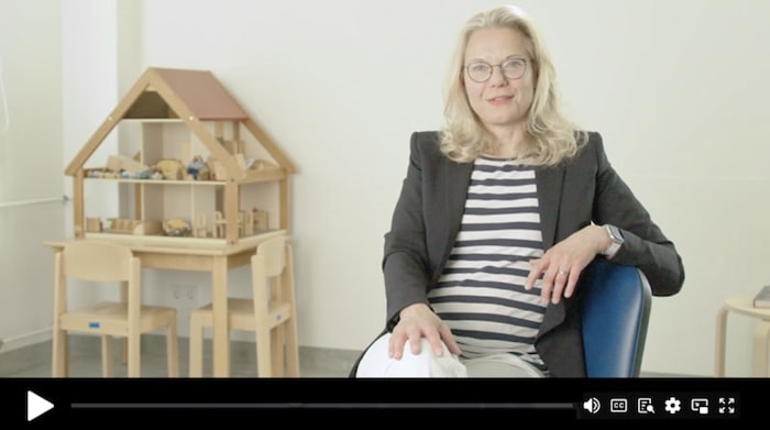 A woman with glasses and blonde hair, wearing a striped shirt and blazer, sits in a room with a wooden dollhouse and chairs in the background.