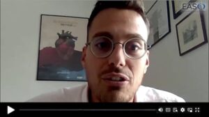 A man wearing round glasses speaks during a video call. The screen displays video controls at the bottom, and there is a movie poster behind him on the wall.