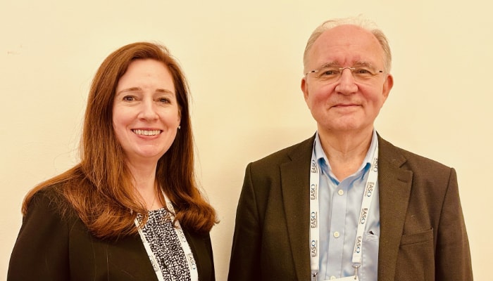 Professor Volkan Yumuk and Dr. Jennifer L. Baker, both wearing conference badges, smiling and standing together in a room with a pale wall.