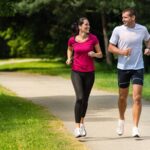 A man and a woman are jogging together on a paved path in a park. They are both smiling as they run, surrounded by green trees and grass.