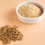 A bowl of mealworm powder sits on a light surface next to a pile of whole mealworms.