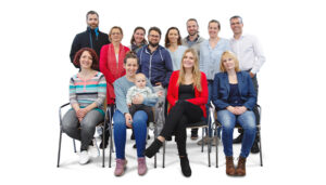 A diverse group of 13 people, including men, women, a baby, and one person holding the baby, posed and seated in two rows against a plain white background.