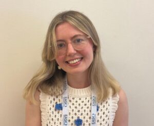 A woman with blonde hair, glasses, and a white knitted top smiles. She wears a lanyard with the text "EASO" printed on it.