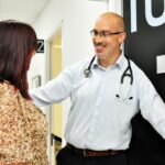 A doctor, with a stethoscope around his neck, standing next to a numbered door, smiles while talking to a female patient in a hallway.